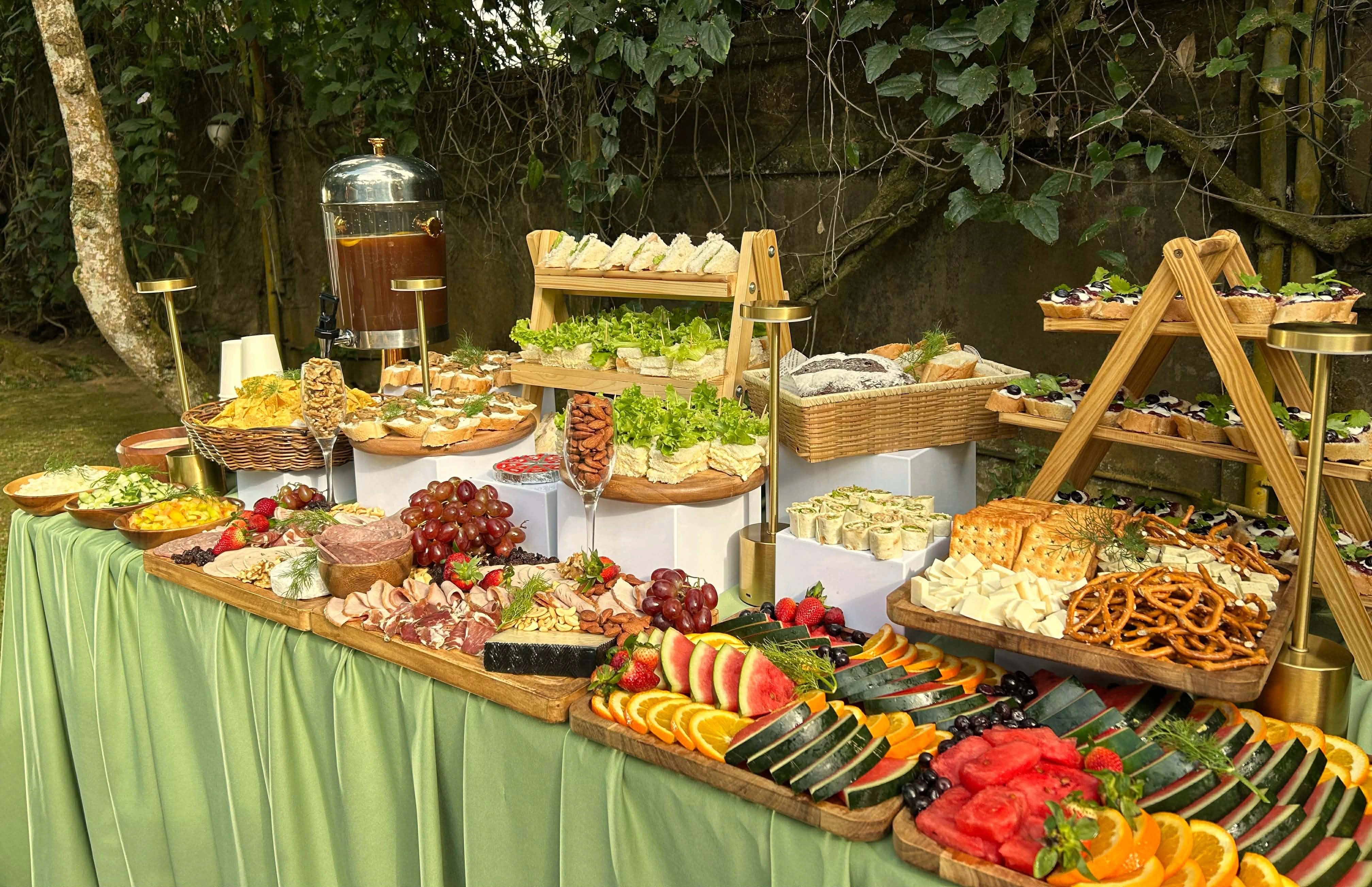 An abundant outdoor grazing table with fruit, charcuterie, cheese, sandwiches, pastries, and refreshments